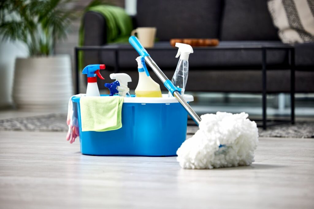 A blue bucket of cleaning supplies on the floor, with a mop, towels, and spray bottles.