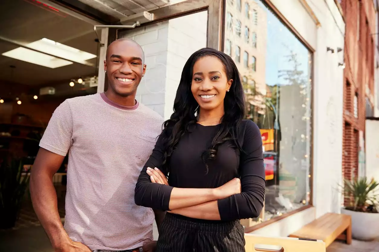 Two young entrepreneurs, a man and a woman, pose and smile outside their small clothing business.